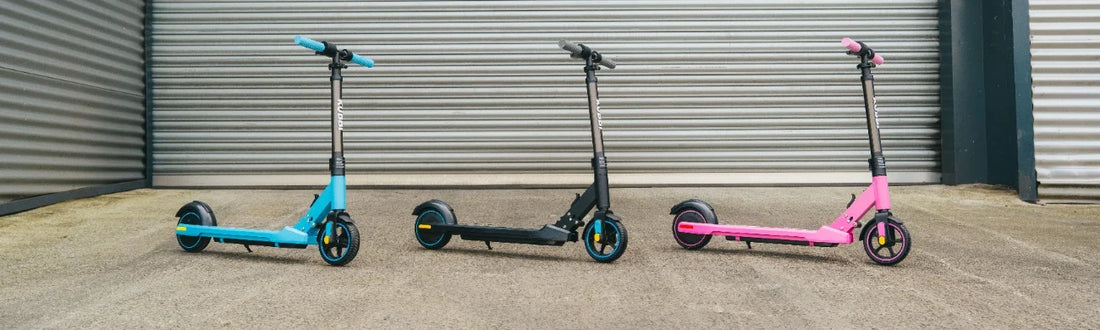 Three Kubbi S4 Pro kids' electric scooters in blue, black and pink, lined up against a shutter