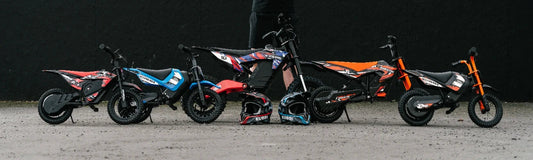 Six Kubbi kids' electric dirt bikes lined up in ascending size against a dark wall, two helmets in front